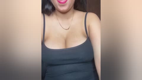 A video of a woman with medium skin tone, wearing a black spaghetti-strap tank top that accentuates her large breasts. Her lips are painted a vibrant pink. She has long, straight black hair. The background is blurred.