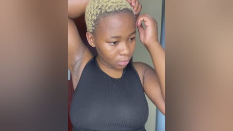 Media: Video of a young Black woman with short, blonde braids, wearing a black ribbed tank top, looking thoughtful while adjusting her hair.