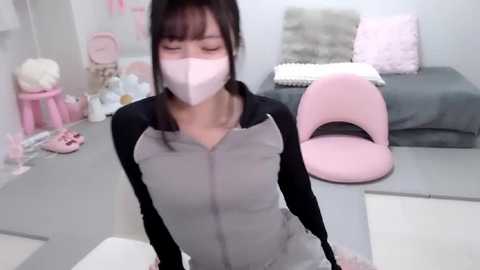 Video of an Asian woman with medium skin tone, wearing a pink face mask, grey and black zip-up hoodie, and grey pants, sitting on a white floor in a pastel-themed room with a pink chair and grey bed.