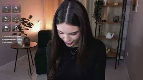 Video of a young woman with long, straight dark hair, wearing a black top and necklace, looking down in a dimly lit, minimalist living room with a green chair, potted plant, and shelves.
