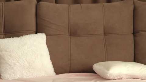A video of a bed with a plush, brown, tufted headboard and a fluffy, white pillow on a pink bedspread.
