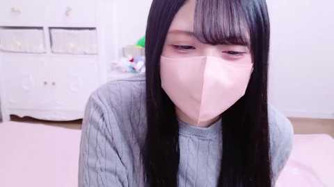 Video of an East Asian woman with long black hair and pale skin, wearing a light gray sweater and a light pink surgical mask. She's indoors, seated on a light pink bed in a minimalist room with white furniture.
