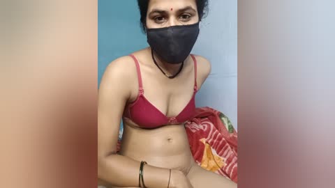 A video of a woman with light brown skin, wearing a red bra and black mask, sitting on a bed with a red and white patterned blanket.
