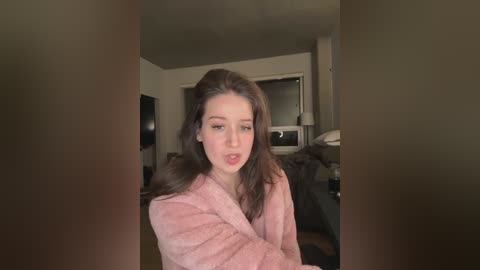 Media: A video of a young woman with long, wavy brown hair, wearing a pink robe, sitting in a dimly lit living room with a TV, couch, and window in the background.