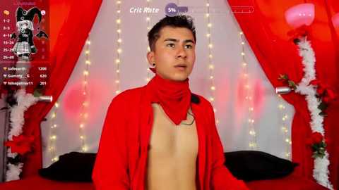 Video of a young, shirtless man with short, dark hair, wearing a red bandana and a red cloak, standing against a backdrop of red drapes and fairy lights.