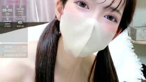 A video of an East Asian woman with long, dark hair in pigtails, wearing a white face mask and a white top, taken indoors with a digital screen displaying live video statistics.