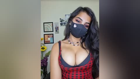 Media: A video of a young woman with long, wavy black hair, wearing a black face mask, red plaid dress, and black bra, standing indoors with framed artwork and sunflowers in the background.