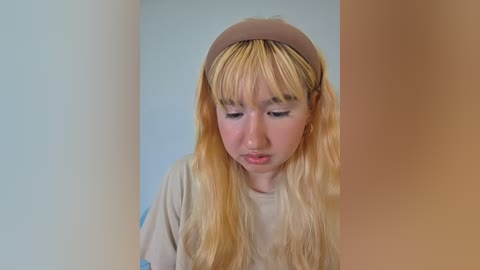 Video of a young woman with long, wavy blonde hair, wearing a beige hat and shirt, standing against a light blue and beige background.