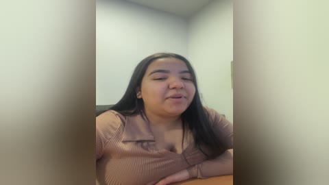 Video of a Latina woman with long black hair, wearing a ribbed, long-sleeved, mauve top, sitting with her eyes closed and mouth slightly open. Background features a plain white wall.