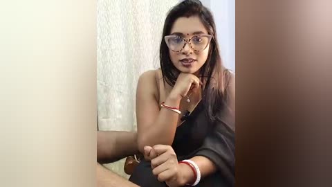 Video of a South Asian woman with medium skin tone, straight dark hair, and clear glasses, wearing a black shirt, seated indoors, holding a phone, with a white textured wall in the background.