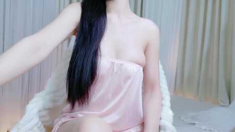 Video of an East Asian woman with long, straight black hair, wearing a pink satin robe, sitting on a white fur blanket in a softly lit room with beige curtains.