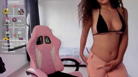Video of a woman with curly hair, wearing a black bikini top and pink shorts, standing in a minimalist room with a pink gaming chair.