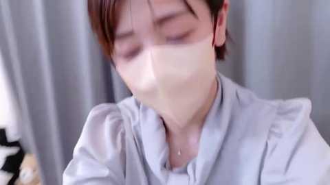 Video of an Asian woman with short brown hair wearing a light blue blouse, with a white surgical mask covering her mouth and nose, looking downward, in a blurred indoor setting with grey curtains.