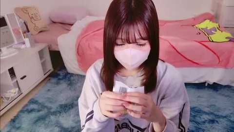 Video of a young Asian woman with straight brown hair, wearing a white mask, white shirt, and blue jeans, sitting on a blue shag rug, playing a handheld video game, in a pink bedroom.