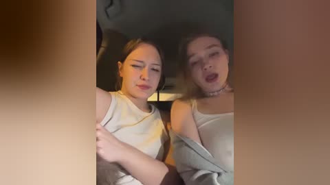 A candid video of two young women, one with light skin and straight hair, the other with fair skin and wavy hair, both smiling and in a relaxed, off-shoulder attire, captured in a dimly lit indoor setting.