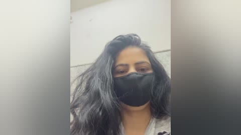 Video of a young woman with long, black hair, wearing a black mask, standing in a dimly lit bathroom with a white wall and a speckled floor.