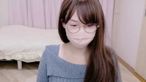 A video of an Asian woman with long brown hair and glasses wearing a blue sweater and face mask, standing in a room with a white bed and pink curtains.