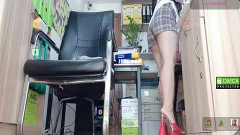 Video of an office with a black leather chair, wooden desk, and a woman in plaid shorts and red heels, leaning forward, with papers and files scattered around.