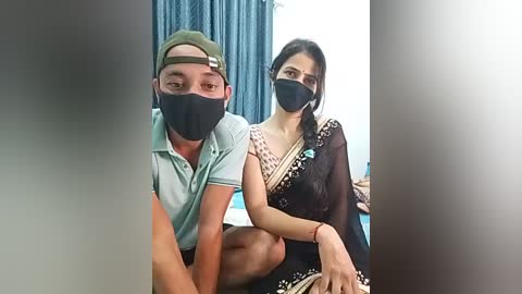 Media: Video of a man and woman wearing masks, sitting closely, in a room with blue curtains and white walls.