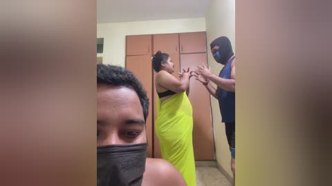 Video of a woman in a bright yellow dress, wearing a black mask, being restrained by a man in a blue shirt, in a dimly lit, beige-walled room.
