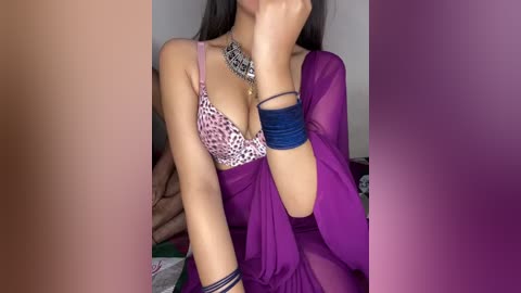 A video of a woman with medium skin tone, wearing a purple sheer robe, leopard print bra, silver necklace, and blue bracelets. Her long dark hair partially covers her face. Background includes a beige couch and a green blanket.