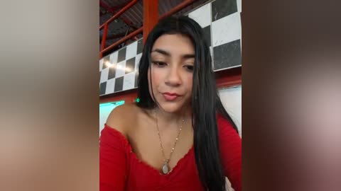 Video of a young Latina woman with long black hair, wearing a red off-shoulder top, standing in a checkered room with a metallic ceiling.