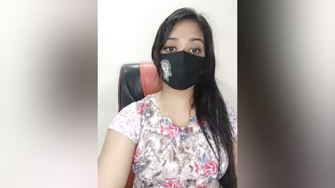 A video of a young woman with long black hair, wearing a floral shirt and black face mask with a white emblem. She sits in a red chair against a blurred background.