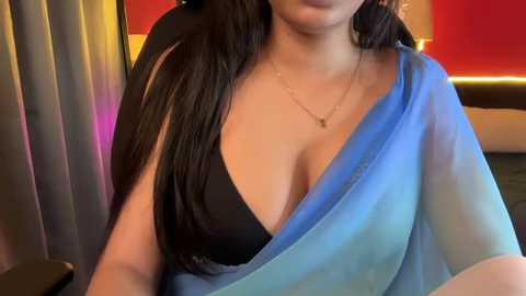 Video of a South Asian woman with long, dark hair, wearing a sheer blue sari over a black bra, a delicate gold necklace, and a blurred indoor setting with red and yellow lighting.