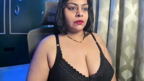 Video of a plus-sized woman with medium skin tone, dark hair, and red lipstick, wearing a black lace bra, sitting in a chair with patterned curtains in the background.