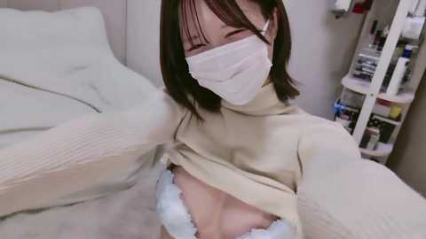 Video of a slender, light-skinned Asian woman with straight brown hair, wearing a white face mask and beige turtleneck sweater, exposing her small breasts with white lace bra. Background includes a white bed and a cluttered medicine cabinet.