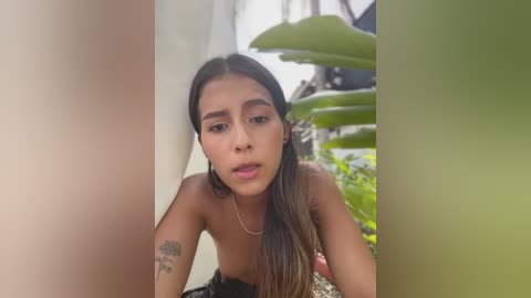 Video of a young, slender, light-skinned Latina woman with long, straight brown hair, topless, wearing black shorts, leaning against a green leafy plant in a garden.