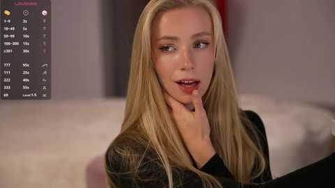 Video of a blonde Caucasian woman with long hair, wearing a black plaid shirt, sitting on a white couch. She touches her chin thoughtfully, with a blurred background featuring a calendar and a wall.