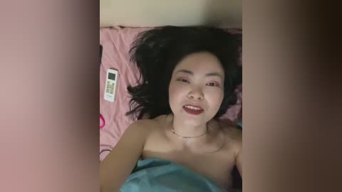 Video of an Asian woman with long black hair, lying on a pink bedspread, smiling, with a remote control beside her.
