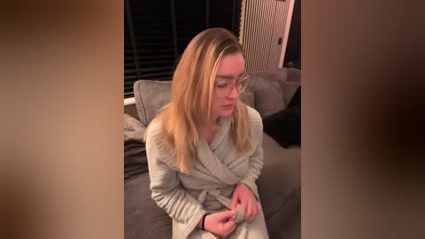 Video of a fair-skinned, blonde woman in a light grey bathrobe, sitting on a grey couch, with a concerned expression, indoors, dim lighting, blinds partially closed.