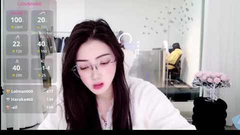 A video of an Asian woman with long black hair, wearing glasses and a white coat, working in a modern office with a digital screen displaying social media metrics.