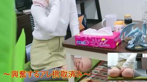 Video of an office desk with tissues, hand sanitizer, a box of crackers, and a white mug. A woman in a white sweater and beige skirt is seated, partially obscured. Japanese text overlay.