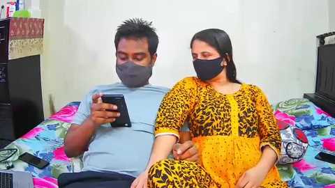Video of a man and woman sitting on a bed with colorful bedding. Man wears a light blue shirt, mask, and phone; woman in yellow dress, mask, and holding phone.