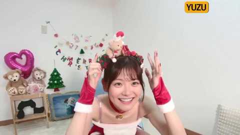 Media: Video of a cheerful Asian woman in a Santa outfit, holding a stuffed bear, in a festive room with holiday decorations and plush toys.