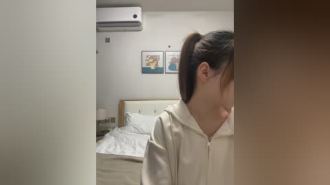 Video of a young woman with light skin and dark hair in a ponytail, wearing a beige hoodie, standing in a modern, minimalist bedroom with a white bed, framed artwork, and an air conditioning unit on the wall.