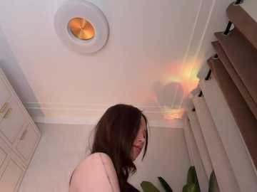 Video of a young woman with long brown hair and glasses, standing in a white-tiled room. She faces right, wearing a light-colored top. A round, glowing light fixture casts rainbow hues on the ceiling.