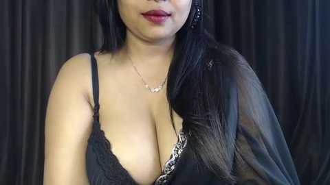 Video of a woman with long black hair, wearing a black lace bra exposing ample cleavage, a delicate necklace, and black sheer robe, against dark curtains.