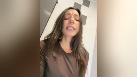 A video of a young woman with long, dark hair, wearing a sheer, dark brown top, leaning against a tiled wall. Her eyes are closed, and she appears to be in a relaxed or possibly intimate moment.