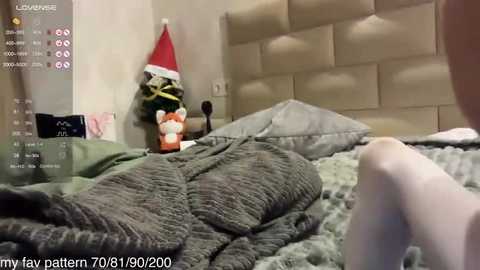 A video of a cozy bedroom featuring a plush, green blanket, a tufted beige headboard, and a red Santa hat on a shelf. A person's legs in white socks are visible in the foreground.