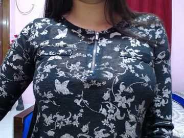 Video of a woman with medium skin tone, wearing a dark blue, long-sleeved, floral-patterned shirt, featuring white flowers and birds. She has long, dark hair and is standing indoors with pink walls and a red curtain in the background.