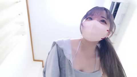Video of an Asian woman with long brown hair, wearing a light grey hoodie and a face mask, standing indoors against a white wall.