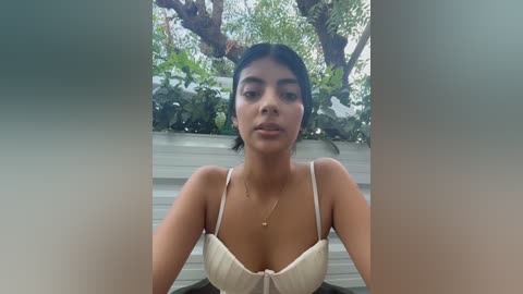 Video of a young Latina woman with medium brown skin, black hair, and a slender build, wearing a beige bra, standing outdoors with lush greenery and a white fence in the background.