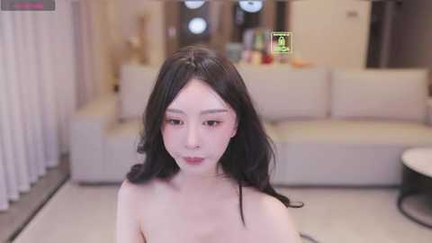 Video of a young Asian woman with long black hair and fair skin, wearing minimal makeup, sitting topless in a modern, minimalist living room with a white sofa and decorative objects.