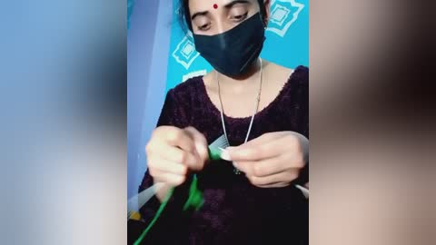 Video of a South Asian woman with a fair complexion, wearing a black mask, maroon sweater, and a bindi, focusing intently on a green object, against a blue wall with white geometric patterns.