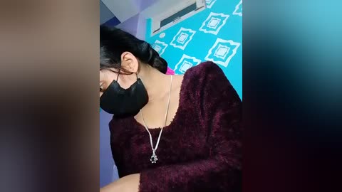 Video of a woman with long black hair, wearing a black face mask, maroon fuzzy sweater, and a silver necklace, looking down, in a room with blue walls adorned with white square patterns.
