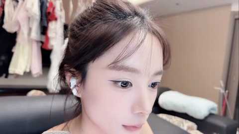 Video of an Asian woman with light skin and dark hair in a bun, wearing white earbuds, looking down, in a beige-walled room with hanging clothes and a black couch.